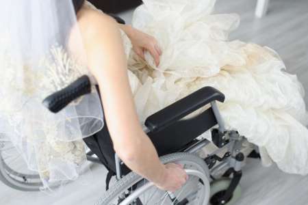 Over the shoulder photo of a white woman pushing her wheelchair with her right arm while wearing a white, lacy veil and bridal gown.